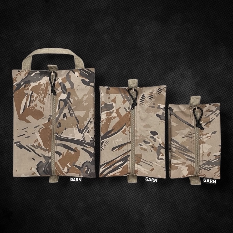 Three arid-camo zip pouches standing side by side in descending sizes on a black textured ground, each with a webbing pull tab, a cord zip pull and a GARN label